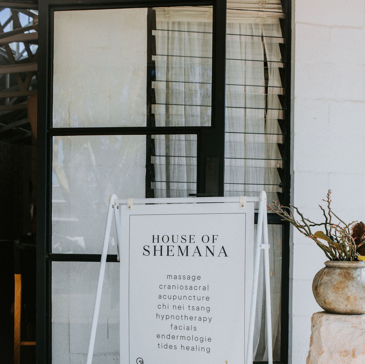 A white A-frame sidewalk sign for 'House of Shemana' stands on a light wooden deck outside a building with black-framed glass windows. A stone planter with dried branches sits to the right.