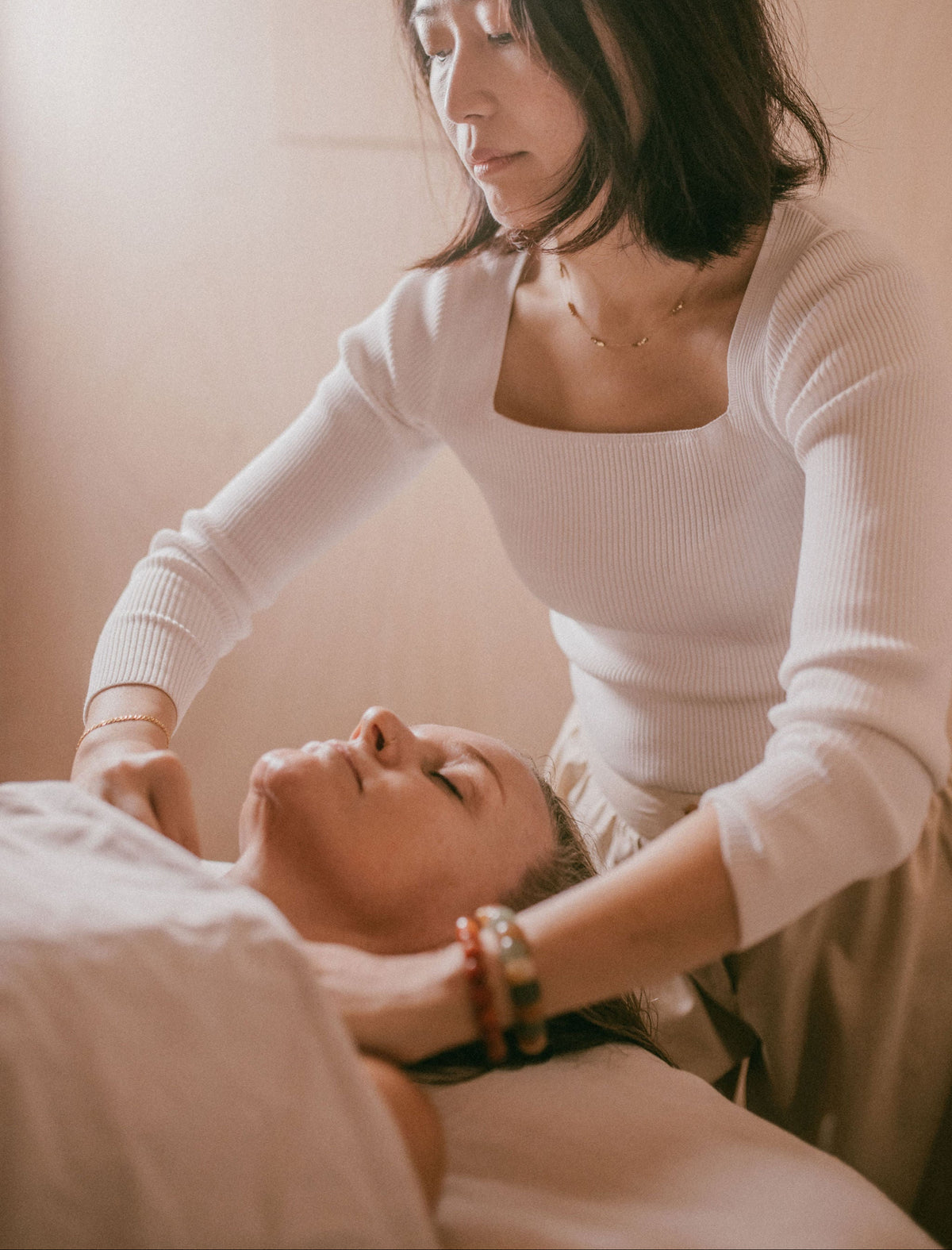 A woman in a white ribbed long-sleeve top performs a head massage on another woman lying down in a spa setting, with warm, soft-focus lighting.