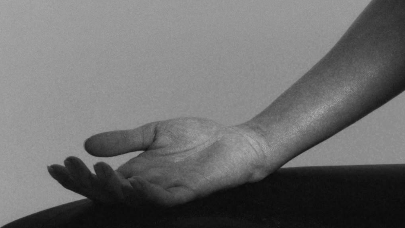 Black and white photograph of a person's hand resting on their knee, with a dark garment covering their lap against a plain light background.